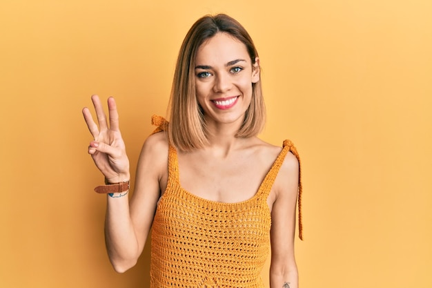 Junge kaukasische blonde Frau trägt ein lässiges gelbes T-Shirt und zeigt mit den Fingern Nummer drei nach oben, während sie selbstbewusst und glücklich lächelt