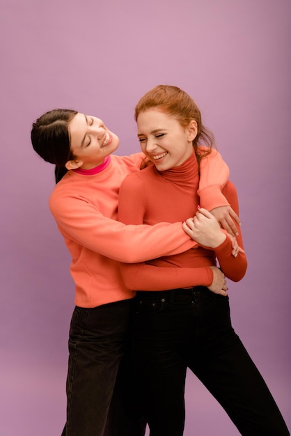 Junge kaukasische brünette Frau in lässiger Kleidung, die ihre lachende rothaarige Freundin von hinten auf violettem Hintergrund umarmt Mood Lifestyle-Konzept