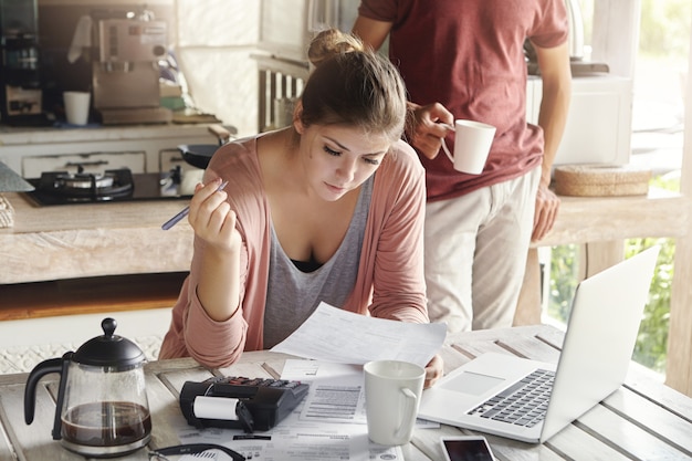 Kostenloses Foto junge kaukasische familie vor finanziellem problem. zufällige frau, die ein stück papier und einen stift hält und dokumente ausfüllt, während sie zahlungen für dienstprogramme leistet, unter verwendung eines taschenrechners und eines allgemeinen laptop-computers