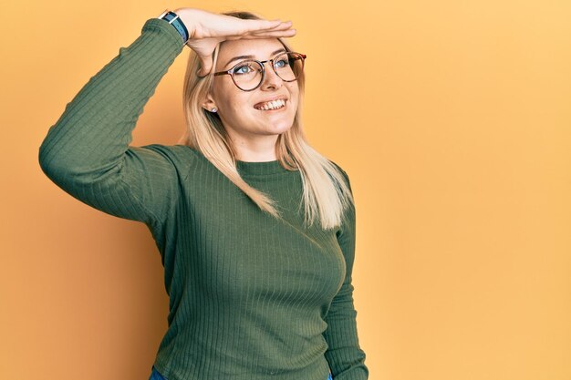 Junge kaukasische Frau in lässiger Kleidung und Brille, sehr glücklich und lächelnd, mit der Hand über dem Kopf in die Ferne schauend und nach einem Konzept suchend