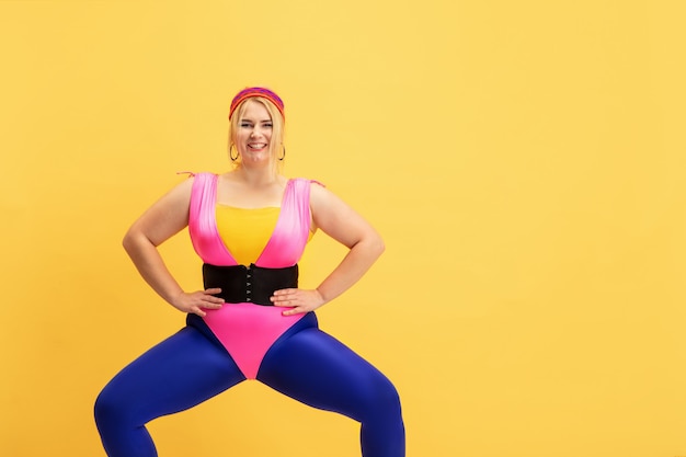 Junge kaukasische plus größe weibliches model training auf gelber wand. exemplar. konzept des sports, gesunder lebensstil, körperpositiv, mode, stil. stilvolle frau, die sich ausdehnt und lächelt.