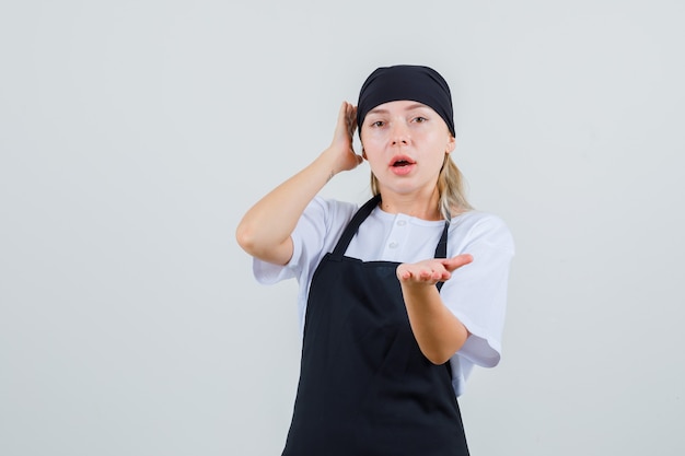 Junge Kellnerin, die Handfläche in fragender Geste in Uniform und Schürze erhebt und verwirrt aussieht