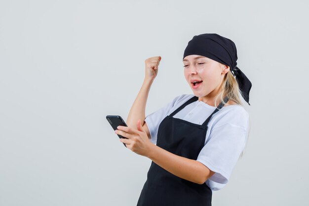 Junge Kellnerin, die Handy in Uniform und Schürze betrachtet und glücklich schaut