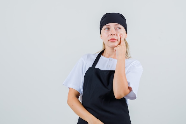 Junge Kellnerin, die mit Finger auf Wange in Uniform und Schürze wegschaut