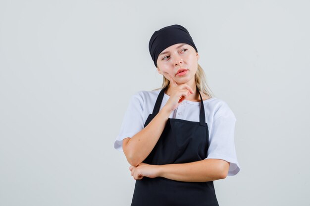 Junge Kellnerin hält Finger auf Wange in Uniform und Schürze und sieht nachdenklich aus