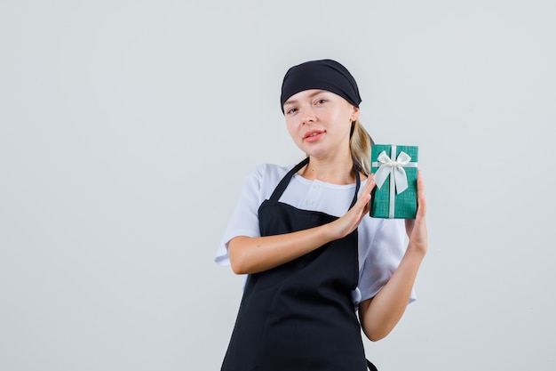 Junge Kellnerin hält Geschenkbox in Uniform und Schürze und sieht positiv aus