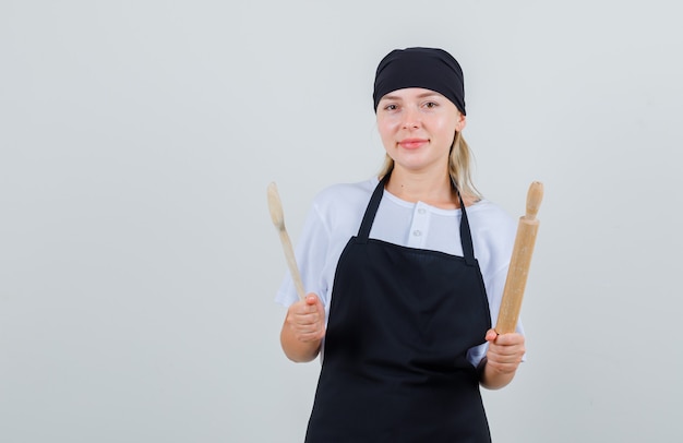 Junge Kellnerin hält Holzlöffel und Nudelholz in Uniform und Schürze und sieht fröhlich aus