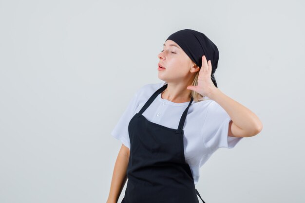 Junge Kellnerin in Uniform und Schürze, die Hand hinter Ohr hält und neugierig schaut