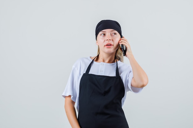 Junge Kellnerin in Uniform und Schürze, die weg schaut, während auf Handy spricht