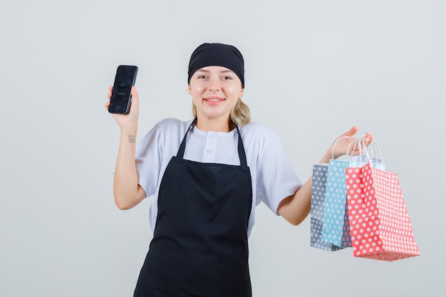 Junge Kellnerin in Uniform und Schürze hält Papiertüten und Handy und sieht fröhlich aus