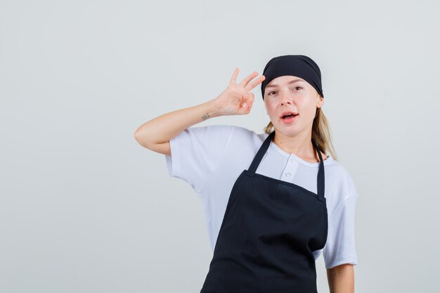 Junge Kellnerin in Uniform und Schürze zeigt ok Zeichen und sieht zufrieden aus