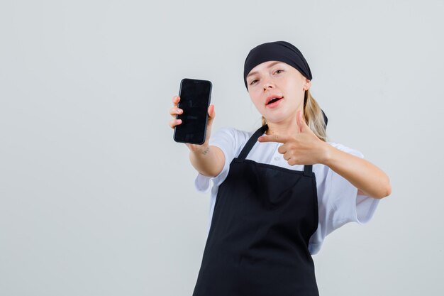 Junge Kellnerin zeigt auf Handy in Uniform und Schürze und sieht zuversichtlich aus