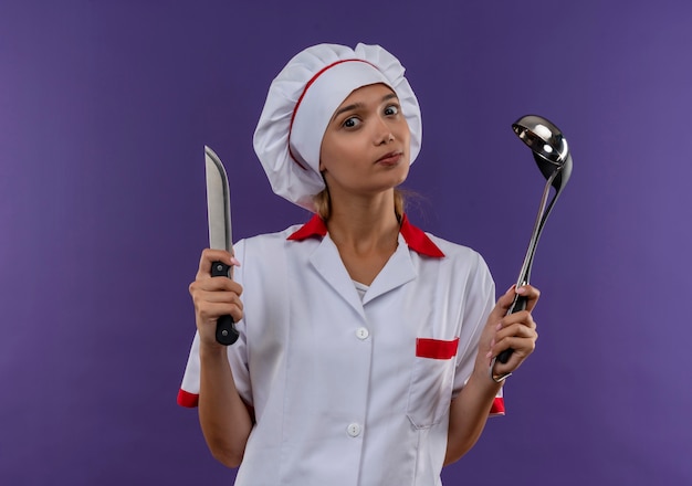 junge Kochfrau, die Kochuniform trägt, die Schöpflöffel und Messer auf isolierter Wand hält