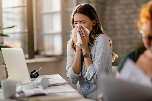 Junge kranke Geschäftsfrau, die während der Arbeit im Büro in ein Taschentuch niest