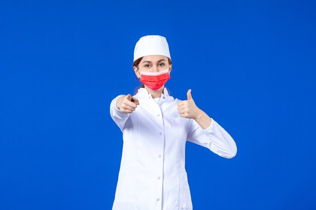 Junge Krankenschwester der Vorderansicht im medizinischen Anzug mit roter Schutzmaske auf blauer Wand