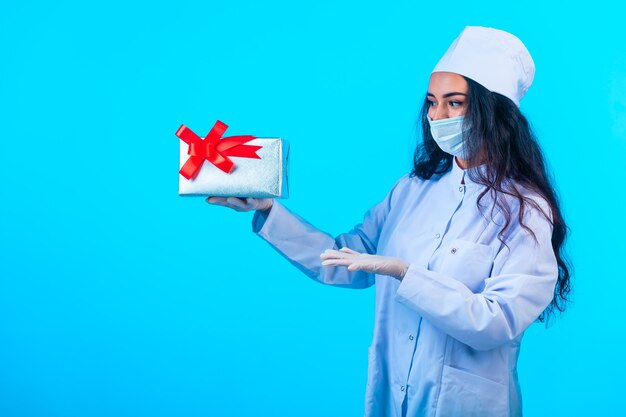 Junge Krankenschwester in isolierter Uniform hält eine Geschenkbox mit rotem Band und präsentiert sie.