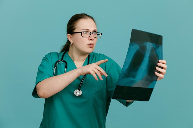 Junge Krankenschwester in medizinischer Uniform mit Stethoskop um den Hals, das Lungenröntgen hält und es sich ansieht, besorgt über blauem Hintergrund zu stehen