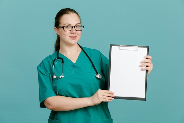 Junge Krankenschwester in medizinischer Uniform mit Stethoskop um den Hals, die ein Klemmbrett hält und in die Kamera blickt, glücklich und positiv lächelnd, selbstbewusst vor blauem Hintergrund