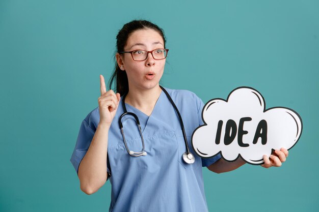 Junge Krankenschwester in medizinischer Uniform mit Stethoskop um den Hals, die eine Sprechblase mit Wortidee hält, die überrascht aussieht und den Zeigefinger zeigt, der eine gute Idee hat, über blauem Hintergrund zu stehen