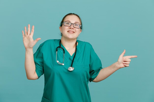 Junge Krankenschwester in medizinischer Uniform mit Stethoskop um den Hals, die fröhlich lächelnd in die Kamera schaut und die Nummer fünf mit offener Handfläche zeigt, die mit dem Zeigefinger zur Seite zeigt