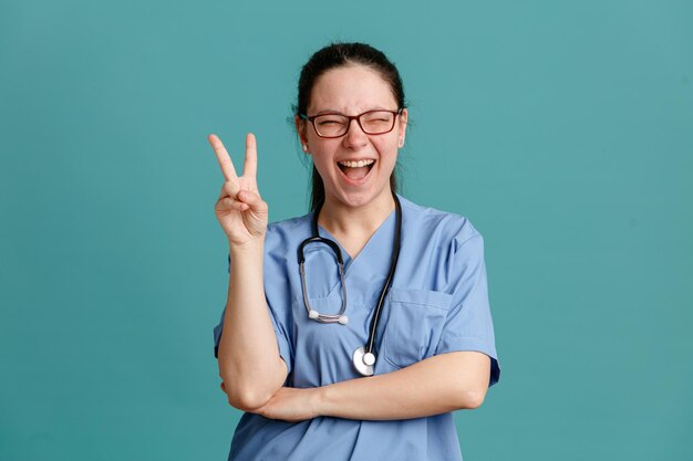 Junge Krankenschwester in medizinischer Uniform mit Stethoskop um den Hals, die glücklich und fröhlich lächelnd in die Kamera schaut und breit Vsign zeigt, das über blauem Hintergrund steht