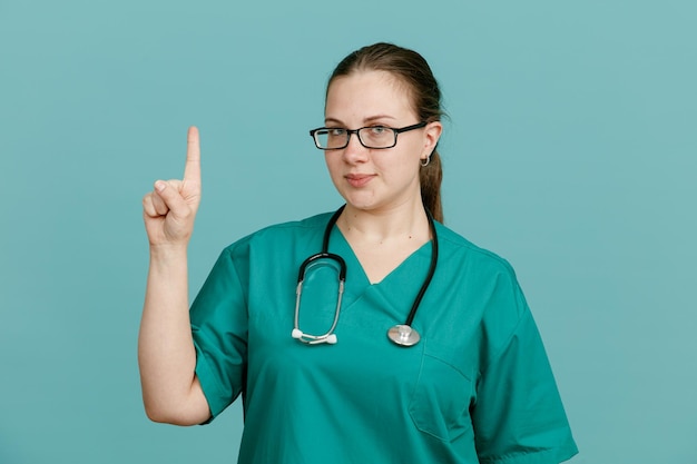 Junge Krankenschwester in medizinischer Uniform mit Stethoskop um den Hals, die in die Kamera blickt und selbstbewusst lächelt und den Zeigefinger zeigt, der eine neue Idee hat, die über blauem Hintergrund steht