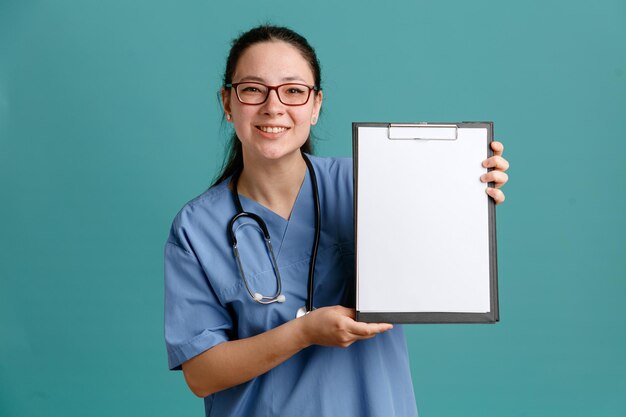 Junge Krankenschwester in medizinischer Uniform mit Stethoskop um den Hals, die Klemmbrett mit leerer Seite hält und in die Kamera schaut, lächelt selbstbewusst, glücklich und positiv über blauem Hintergrund