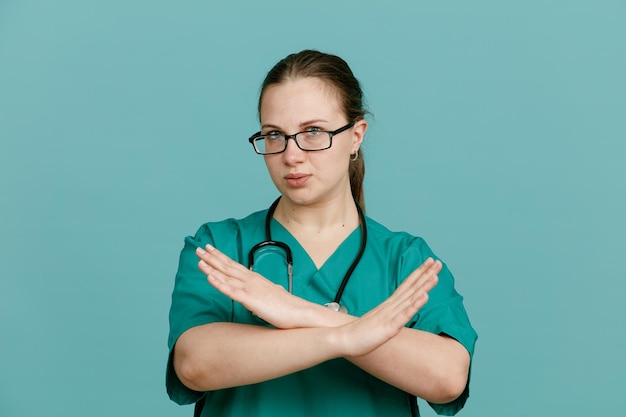 Junge Krankenschwester in medizinischer Uniform mit Stethoskop um den Hals, die mit ernstem Gesicht in die Kamera schaut und eine Stoppgeste macht, die ihre Hände über blauem Hintergrund kreuzt