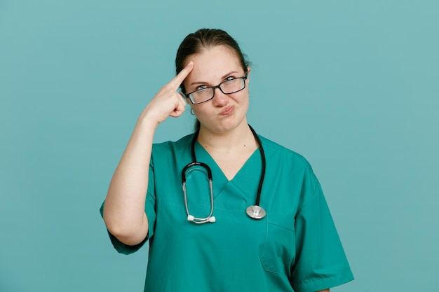 Junge Krankenschwester in medizinischer Uniform mit Stethoskop um den Hals, die verwirrt in die Kamera blickt und mit dem Zeigefinger auf ihre Schläfe zeigt, weil sie über blauem Hintergrund steht