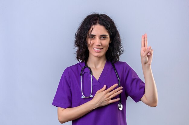 Junge Krankenschwester in medizinischer Uniform und mit dem Stethoskop, das die Hand hebt, um Eide zu leisten, versprechen, nur die Wahrheit zu sagen, seien Sie aufrichtig und ehrlich und schauen Sie mit einem selbstbewussten Lächeln über Isolat