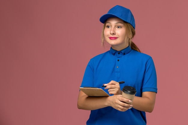 Kostenloses Foto junge kurierin der vorderansicht in der blauen uniform, die das halten der tasse kaffee und des notizblocks mit leichtem lächeln aufwirft, dienstuniformenlieferungsarbeiterin