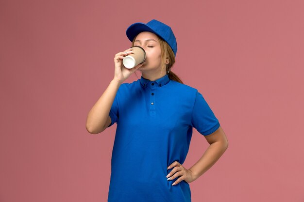 Junge Kurierin der Vorderansicht in der blauen Uniform, die deivery Kaffee auf der rosa Wand, Dienstjobuniform-Lieferfrau aufwirft und trinkt