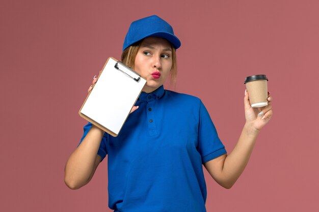 Junge Kurierin der Vorderansicht in der blauen Uniform, die die Tasse Kaffee und den Notizblock hält, Dienstuniform-Lieferarbeiter hält