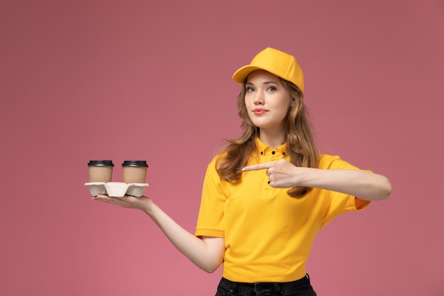 Junge Kurierin der Vorderansicht in der gelben Uniform, die braune Kaffeetassen des Plastiks braun auf dem rosa Schreibtischjobuniformlieferfarbdienstarbeiter hält