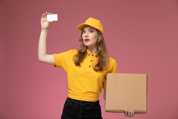 Junge Kurierin der Vorderansicht in der gelben Uniform, die braune Nahrungsmittelbox mit weißer Karte auf dem weiblichen Arbeiter des dunkelrosa Schreibtischuniformlieferdienstes hält