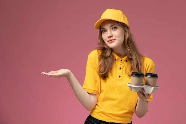 Junge Kurierin der Vorderansicht in der gelben Uniform, die Kaffeetassen auf dunkelrosa Schreibtischuniformlieferdienstarbeiter hält