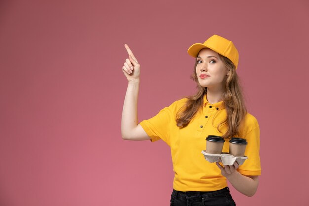 Junge Kurierin der Vorderansicht in der gelben Uniform, die Kaffeetassen hält, die auf dunkelrosa Schreibtischuniformlieferdienstarbeitsarbeiter aufwirft