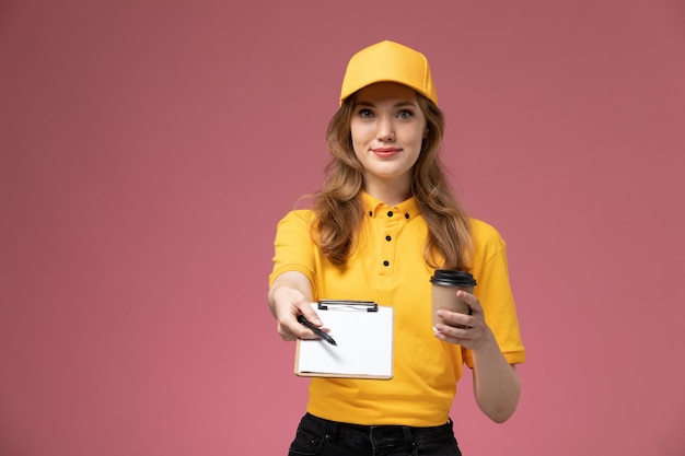 Junge Kurierin der Vorderansicht in der gelben Uniform, die Plastikkaffeetasse mit Notizblock auf dem dunkelrosa Schreibtischuniformlieferdienstarbeitsarbeiter hält