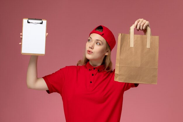 Junge Kurierin der Vorderansicht in der roten Uniform, die Lieferung-Nahrungsmittelpaket und Notizblock auf der hellrosa Wand hält