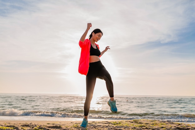 Junge lächelnde attraktive schlanke Frau, die Sport im Morgensonnenaufgang macht, der auf Meeresstrand in Sportkleidung, gesundem Lebensstil springt, Musik auf Kopfhörern hörend, rosa Windjacke tragend, Spaß habend