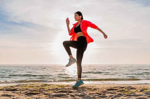 Junge lächelnde attraktive schlanke Frau, die Sport im Morgensonnenaufgang macht, der auf Meeresstrand in Sportkleidung, gesundem Lebensstil springt, Musik auf Kopfhörern hörend, rosa Windjacke tragend, Spaß habend