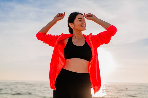 Junge lächelnde attraktive schlanke Frau, die Sport im Morgensonnenaufgang tanzt, der auf Meeresstrand in Sportkleidung, gesundem Lebensstil tanzt, Musik auf Kopfhörern hörend, rosa Windjacke tragend, Spaß habend