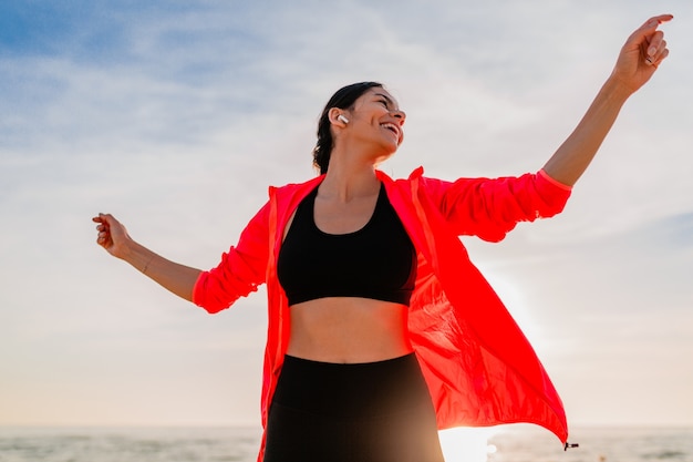 Junge lächelnde attraktive schlanke Frau, die Sport im Morgensonnenaufgang tanzt, der auf Meeresstrand in Sportkleidung, gesundem Lebensstil tanzt, Musik auf Kopfhörern hörend, rosa Windjacke tragend, Spaß habend