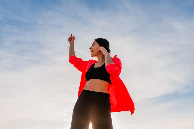 Junge lächelnde attraktive schlanke Frau, die Sport im Morgensonnenaufgang tanzt, der auf Meeresstrand in Sportkleidung, gesundem Lebensstil tanzt, Musik auf Kopfhörern hörend, rosa Windjacke tragend, Spaß habend