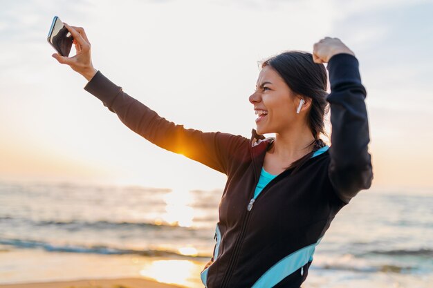 Junge lächelnde attraktive schlanke Frau, die Sportübungen am Morgensonnenaufgangstrand in Sportbekleidung, gesundem Lebensstil, Musik auf Kopfhörern hörend macht, Selfie-Foto auf Telefon schauend stark macht