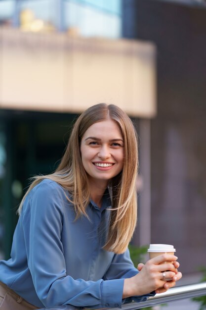Junge lächelnde berufstätige Frau, die eine Kaffeepause während ihres vollen Arbeitstages hat. Sie hält einen Pappbecher im Freien in der Nähe des Geschäftsgebäudes, während sie sich entspannt und ihr Getränk genießt.