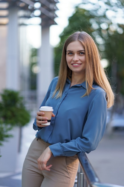 Junge lächelnde berufstätige Frau, die eine Kaffeepause während ihres vollen Arbeitstages hat. Sie hält einen Pappbecher im Freien in der Nähe des Geschäftsgebäudes, während sie sich entspannt und ihr Getränk genießt.