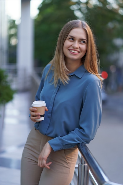 Junge lächelnde berufstätige frau, die eine kaffeepause während ihres vollen arbeitstages hat. sie hält einen pappbecher im freien in der nähe des geschäftsgebäudes, während sie sich entspannt und ihr getränk genießt.