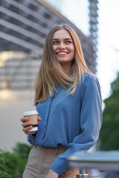 Junge lächelnde berufstätige Frau, die eine Kaffeepause während ihres vollen Arbeitstages hat. Sie hält einen Pappbecher im Freien in der Nähe des Geschäftsgebäudes, während sie sich entspannt und ihr Getränk genießt.