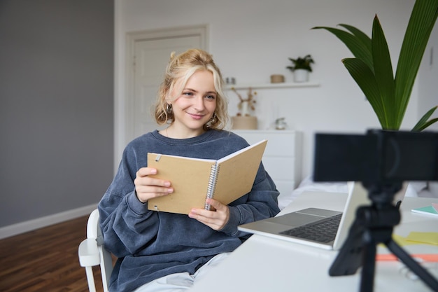 Junge lächelnde blonde Frau sitzt in der Nähe eines Laptops, benutzt eine digitale Kamera, um Videos aufzunehmen, erstellt einen Blog